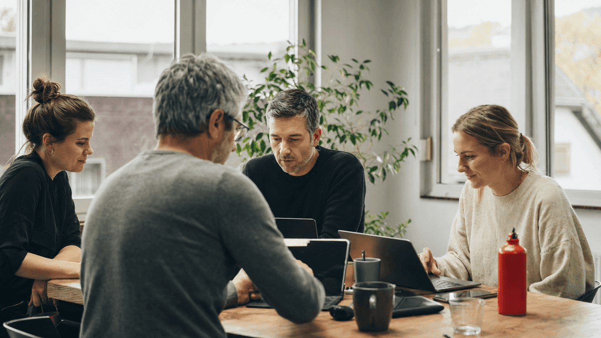 A small team of four employees in a typical Austrian SME office, mix of ages, gathered around a table with laptops and coffee, casual workday atmosphere, daylight through windows, authentic and photorealistic, no visible text