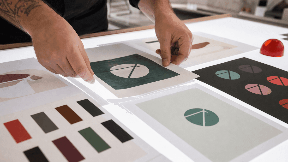 Close-up of a graphic designer's hands arranging printed brand identity sheets on a white lightbox table — logo variants, color palette chips, and typeface samples fanned out in an organized layout, soft studio lighting, no readable text