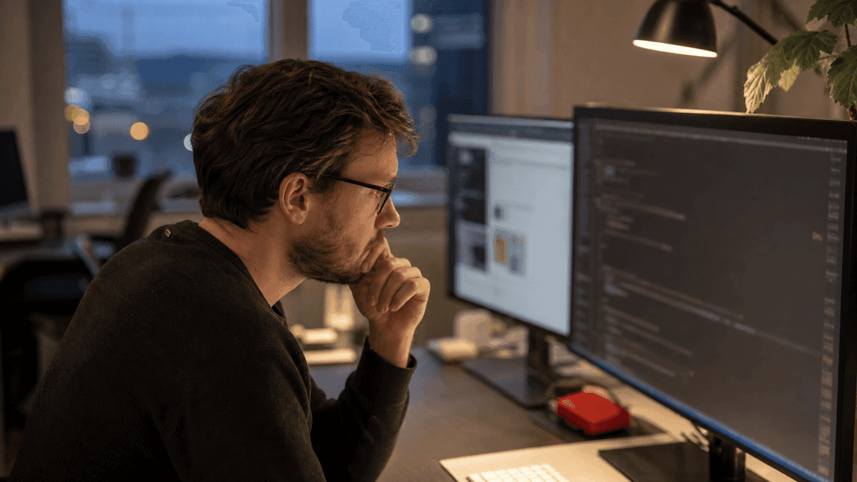 A concentrated developer at a desk closely reviewing code on a large monitor, brow slightly furrowed while spotting a subtle error, second screen with a website preview, evening office light, authentic and photorealistic, no readable text