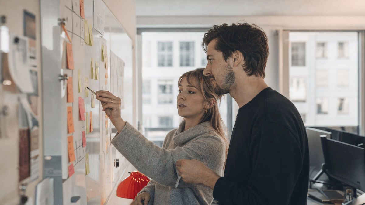 Two marketing colleagues collaborating at a whiteboard with sticky notes and a step-by-step action plan, natural daylight in a modern Vienna office, authentic teamwork moment, photorealistic, no readable text